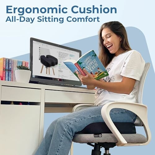 Femme souriante assise sur un coussin ergonomique en lisant un livre devant un ordinateur.